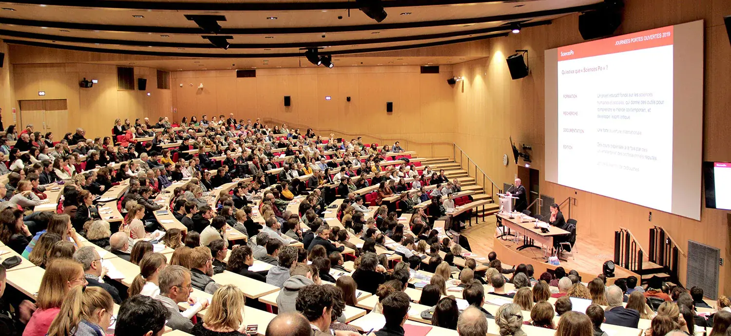 Amphithéatre où des centaines d'étudiants écoutent deux professeurs parlés sur leur estrade.