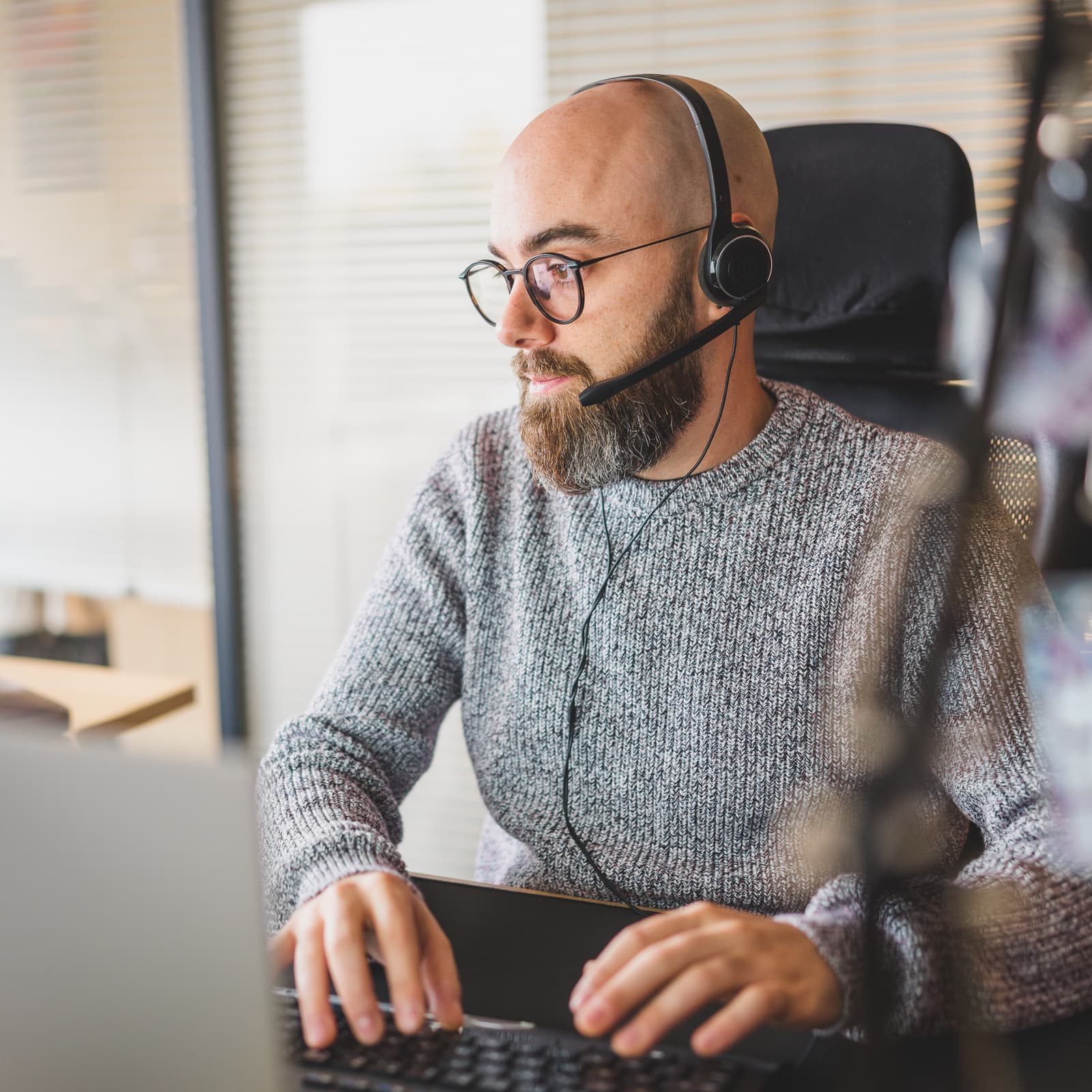 Homme à lunette portant un casque audio qui regarde son ordinateur et qui tape sur les touches de son clavier.