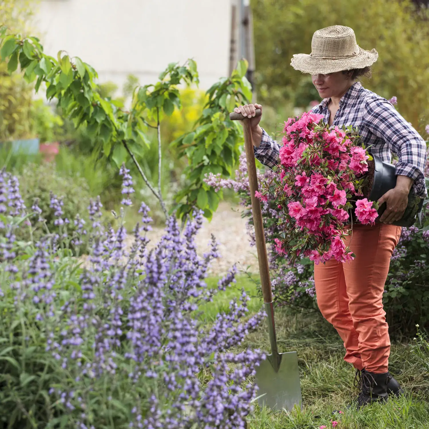 Femme avec un chapeau portant dans son bras un pot de fleurs, qui jardine dans un jardin à l'extérieur et qui à une pelle dans son autre main.