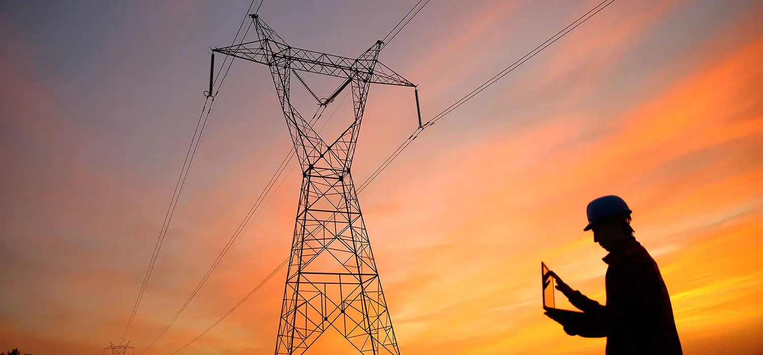 Pylône électrique dans un champ où un homme avec un casque de chantier est en train de vérifier des éléments sur son ordinateur.