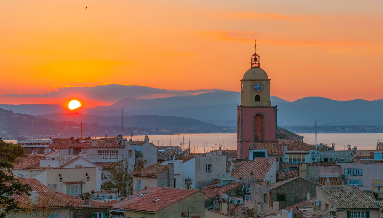 Coucher de soleil flamboyant sur les toits provençaux de Saint-Tropez, mettant en valeur son célèbre clocher rose et jaune face à la mer et aux montagnes.