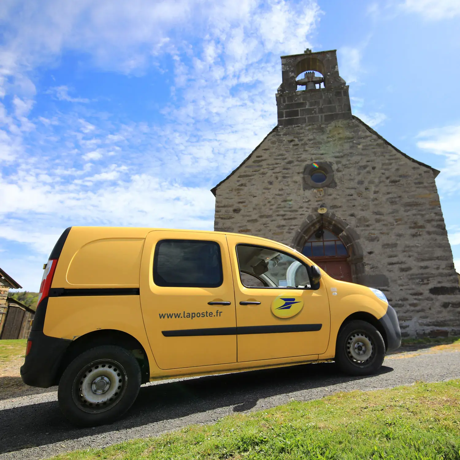 Voiture jaune floqué avec le logo de La Poste garée devant une chapelle.